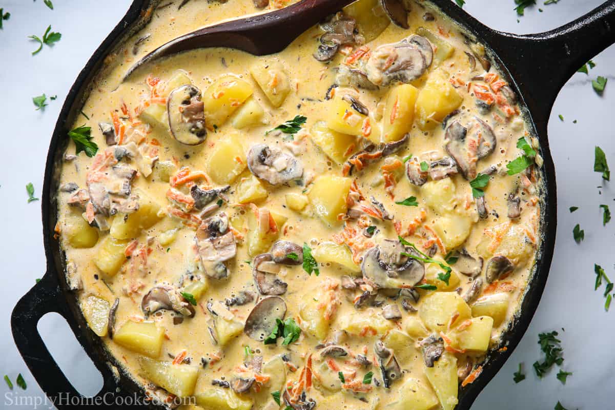 Skillet full of Potatoes with Mushroom Gravy on a white background with a wooden spoon stirring it.