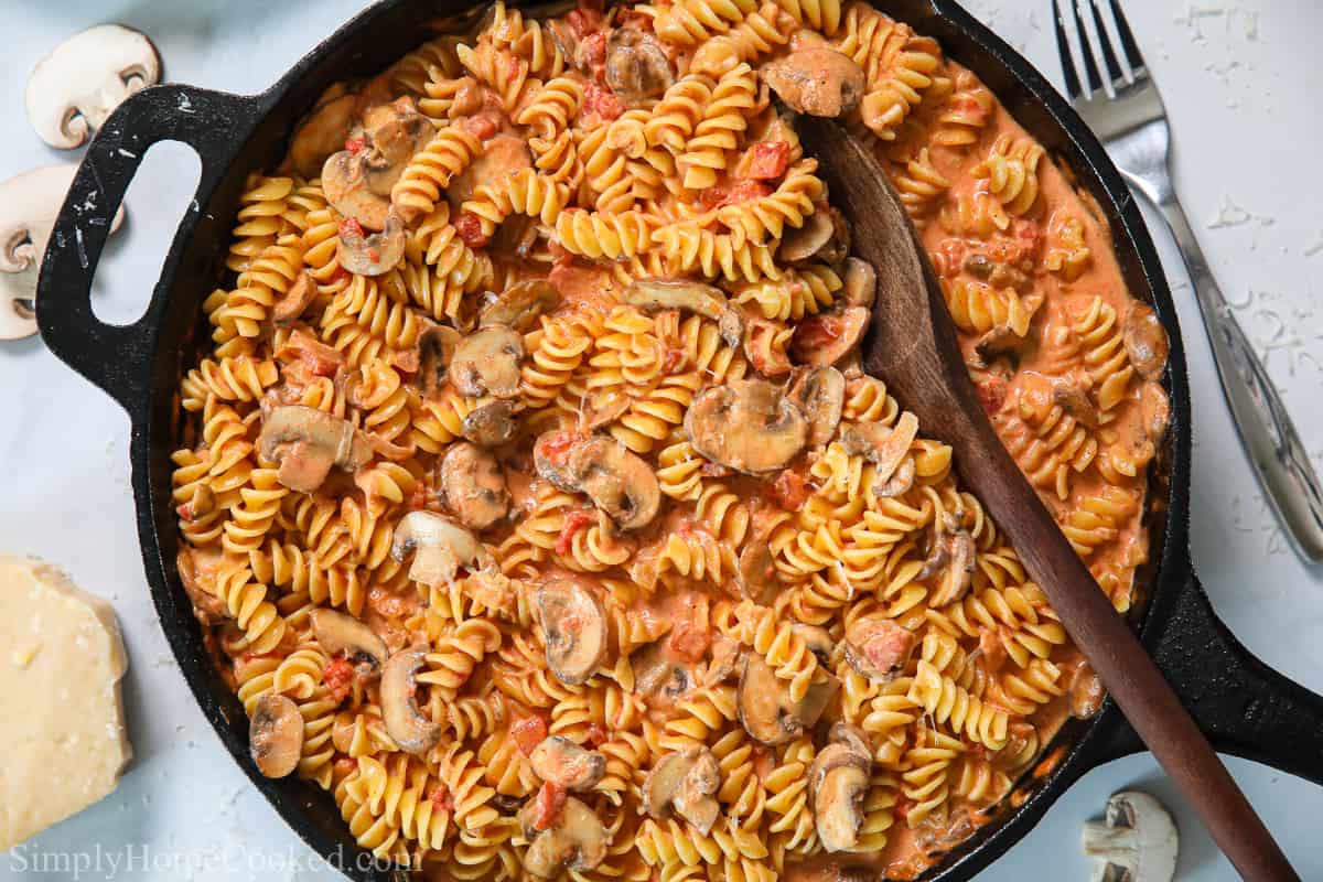 Skillet of Creamy Tomato and Mushroom Rotini Pasta with a wooden spoon in it and some sliced mushrooms, Parmesan, and a fork in the background.