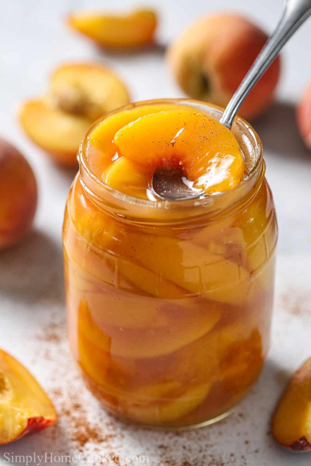 Glass jar with Peach Pie Filling and a spoon scooping some out, with peaches in the white background.