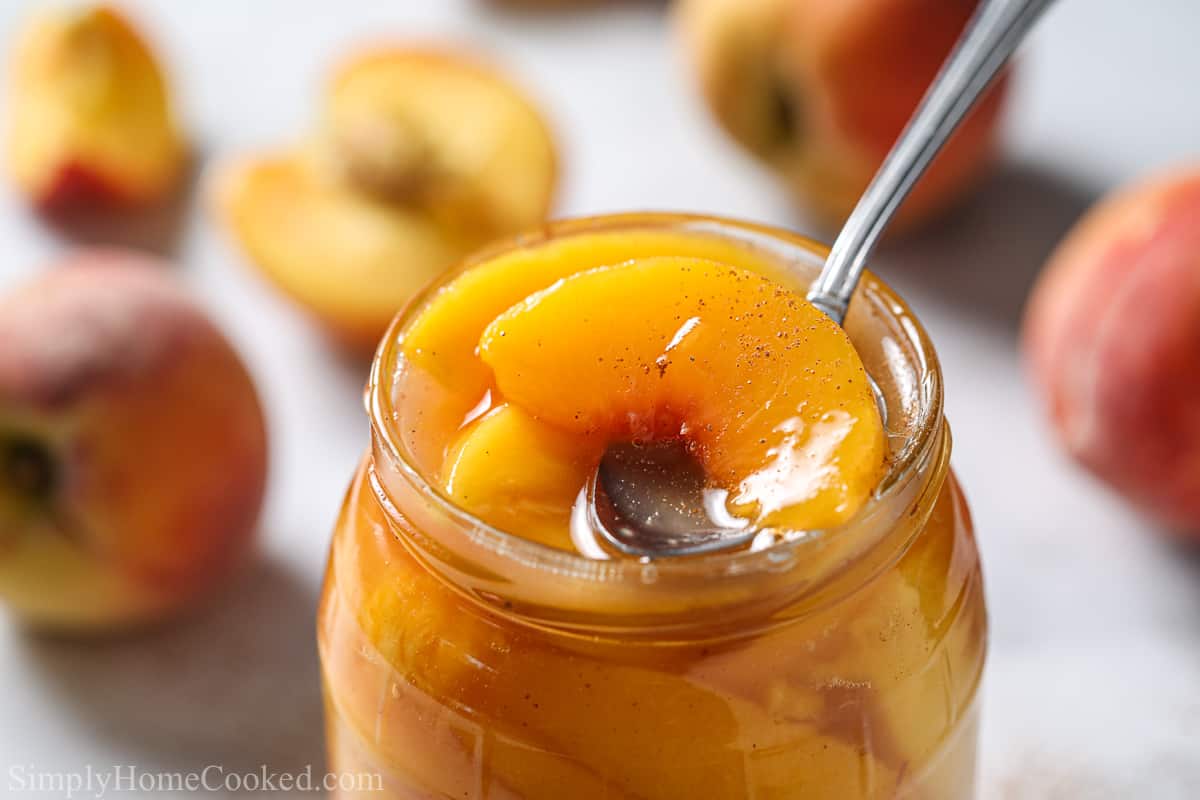 Close up of glass jar with Peach Pie Filling and a spoon scooping some out, with peaches in the white background.