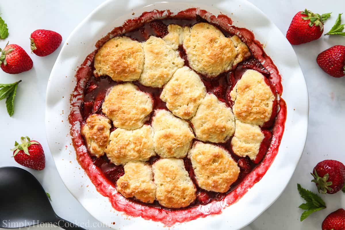 Strawberry Fruit Cobbler in a white pie dish with strawberries and a serving spoon in the background.