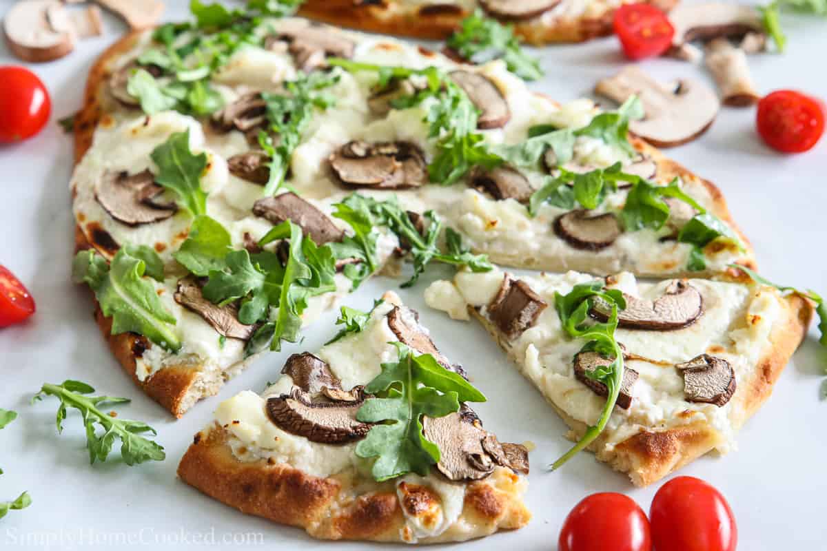 Close up of Mushroom Arugula Flatbread Pizza, sliced, with cherry tomatoes and other ingredients scattered on a white background.