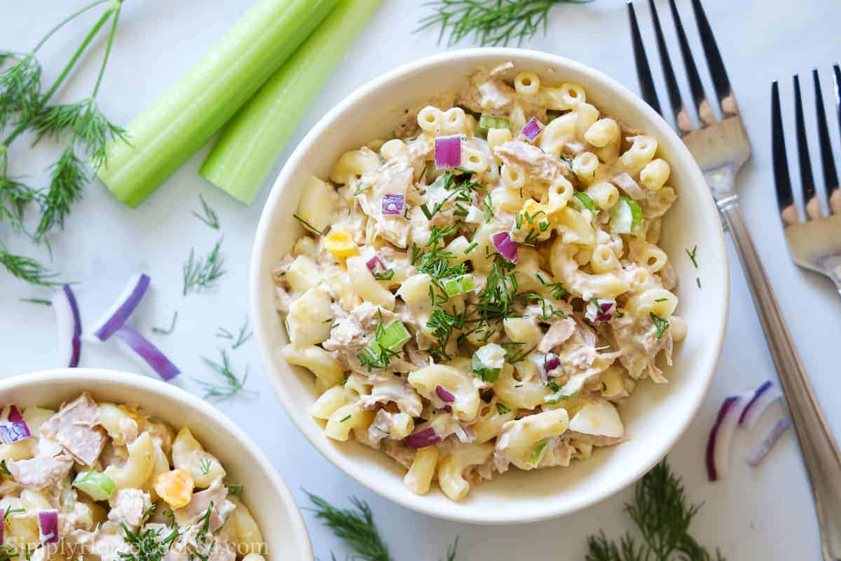 2 bowls of Tuna Pasta Salad with forks, celery, onion, and dill scattered nearby.