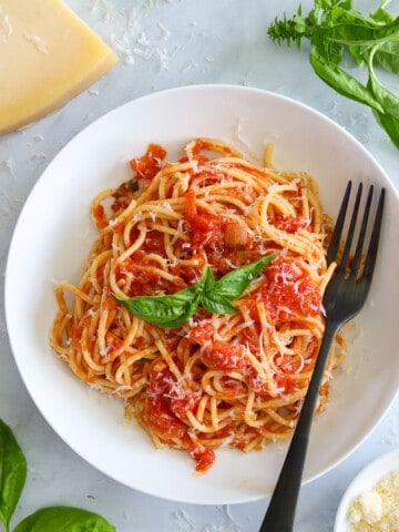 Pasta Pomodoro on a white plate with a fork and Parmesan cheese and basil nearby.