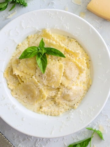 White plate of Easy Four Cheese Ravioli with herbs on top and some herbs and Parmesan cheese to the side.