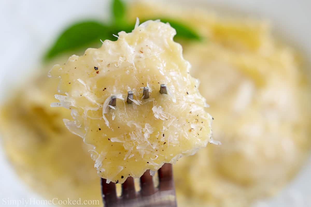 Close up of a fork with a Four Cheese Ravioli on it, a plate of ravioli in the background.