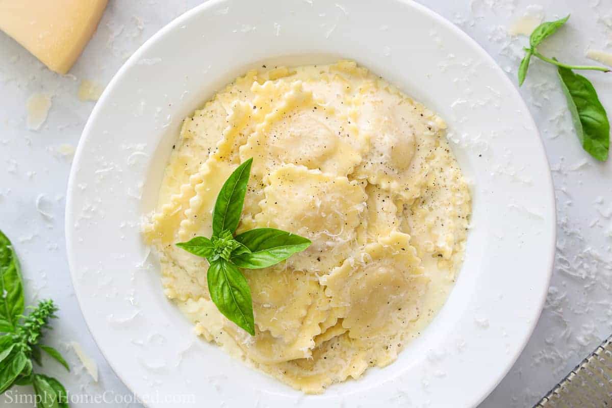 Overhead view of a white plate full of Easy Four Cheese Ravioli in a cream sauce with herbs on top and to the side.