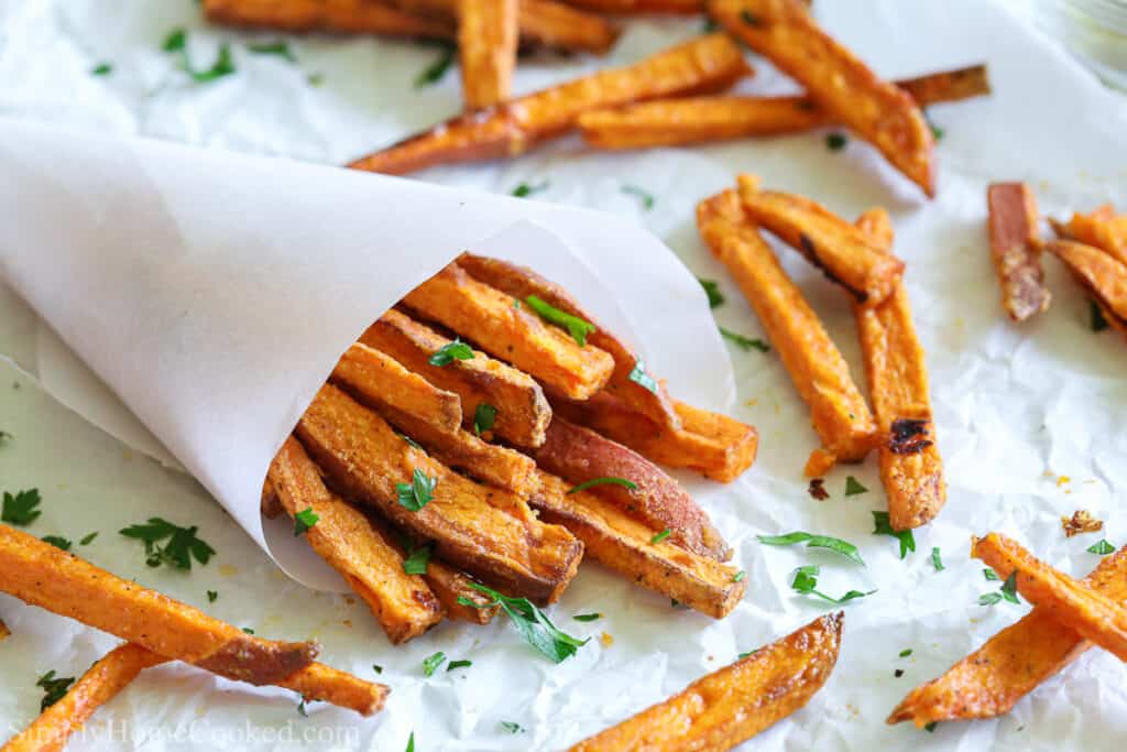 Paper cone of the BEST Air Fryer Sweet Potato Fries, with other fries and parsley scattered on a white paper background.