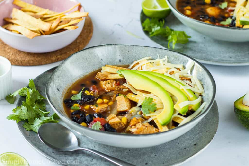 Bowl of Chicken Tortilla Soup with  avocado, cheese, and tortilla strips on top, cilantro and a spoon on the side, and a bowl of tortilla strips and more soup in the background.