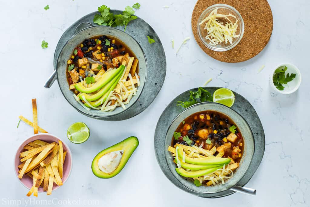 2 bowls of Chicken Tortilla Soup with  avocado, cheese, and tortilla strips on top, lime halves and cilantro on the side, and an avocado, lime, cheese, cilantro, and tortilla strips nearby.