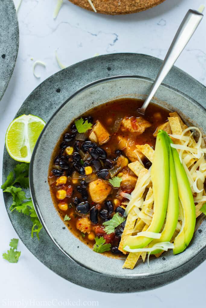 Close up of a bowl of Chicken Tortilla Soup with  avocado, cheese, and tortilla strips on top and s poon in it; lime and cilantro on the side.