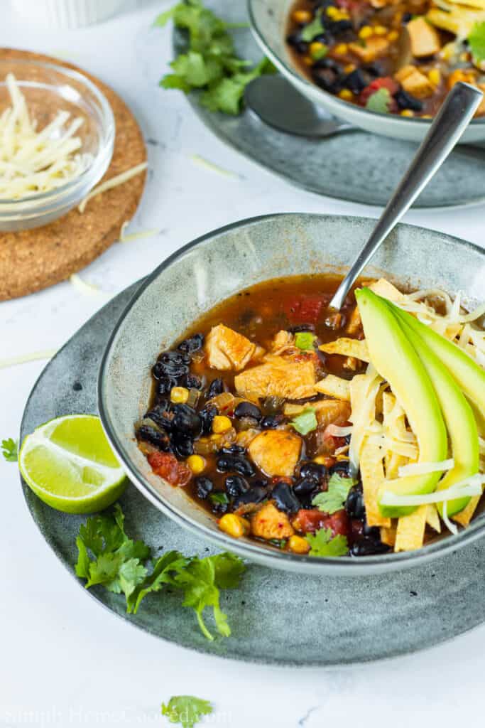Bowl of Chicken Tortilla Soup with a spoon in it, avocado, cheese, and tortilla strips on top, a lime on the side, and other ingredients in the background.