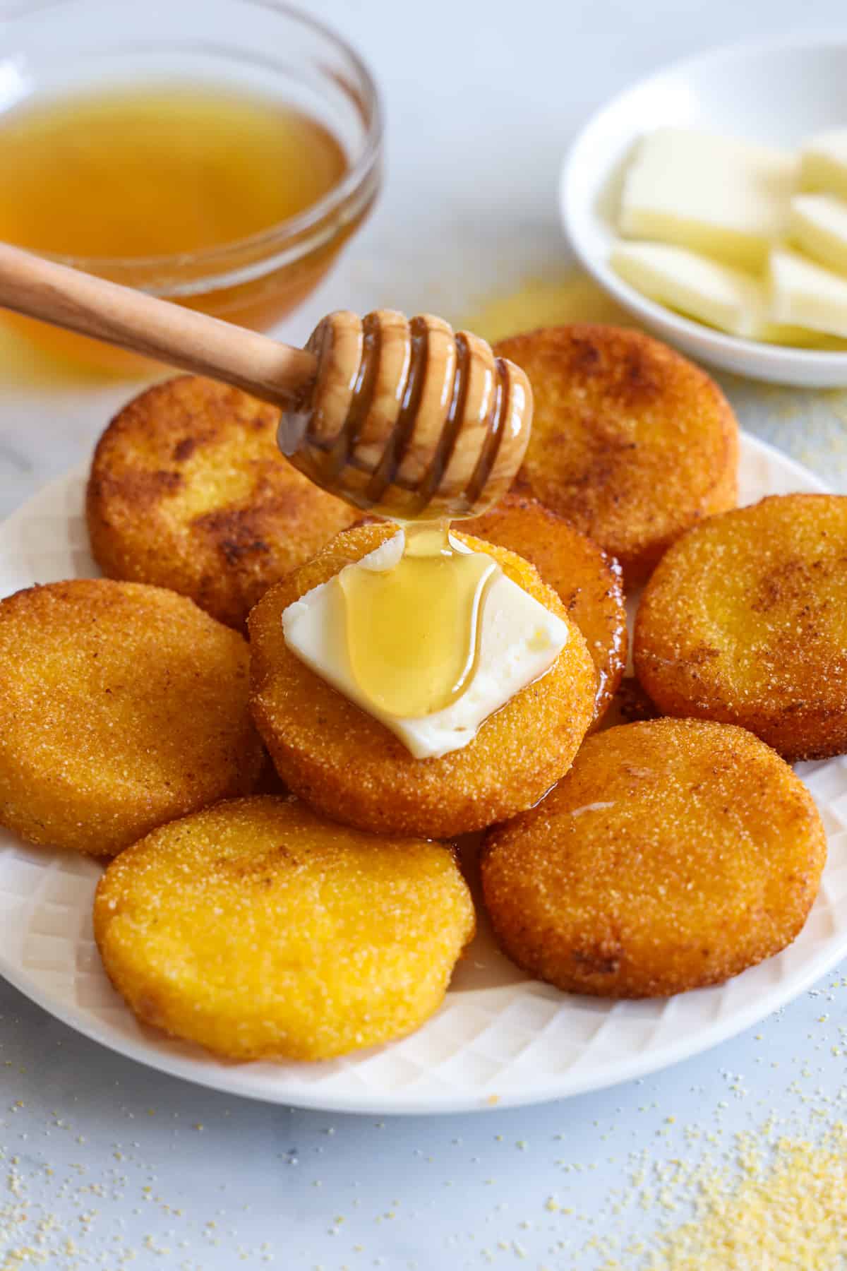 Southern Hot Water Cornbread on a plate with a pat of butter and honey being dribbled on it, honey and butter in the background.