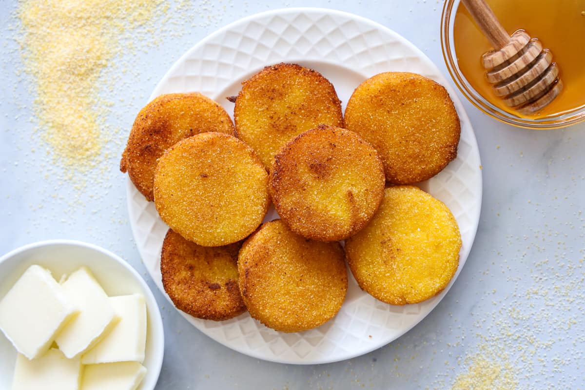 Vista dall'alto del sud Cornbread acqua calda su un piatto bianco con una ciotola di miele e un altro di burro nelle vicinanze, alcuni farina di mais sparsi a lato.