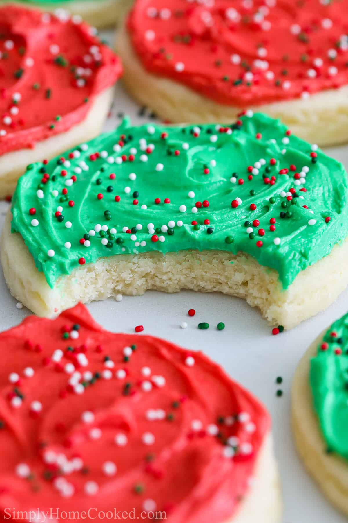 Vertical image of a Close up of Lofthouse Frosted Sugar Cookies with red and green frosting and sprinkles, one with a bite taken out of it.