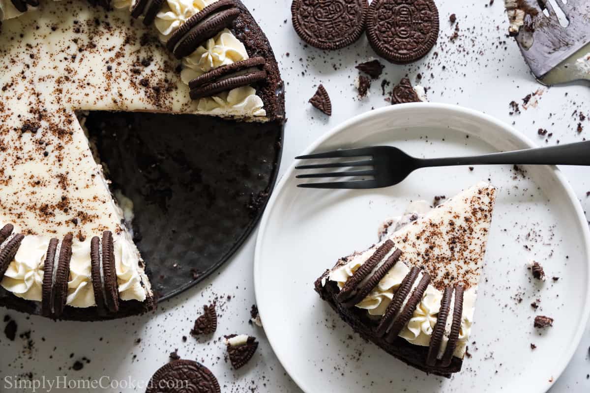 Aerial view of Easy Oreo Cheesecake with slide missing and a white plate with a fork and slice of cheesecake, Oreos and crumbs in the background.