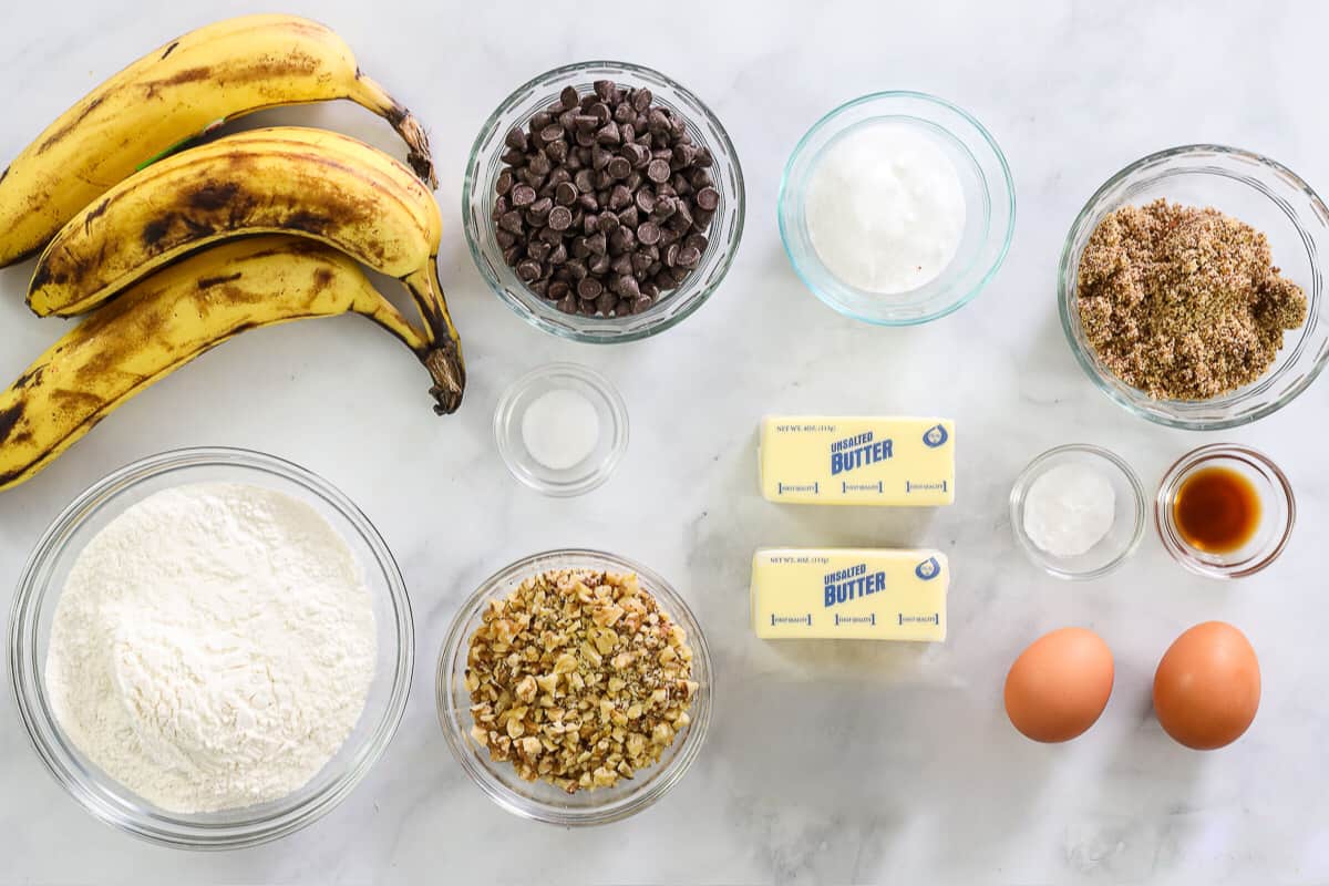 Ingredients for Banana Chocolate Chip Muffins, including bananas, chocolate chips, butter, sugar, brown sugar, eggs, walnuts, flour, salt, baking soda, and vanilla, on a white background.