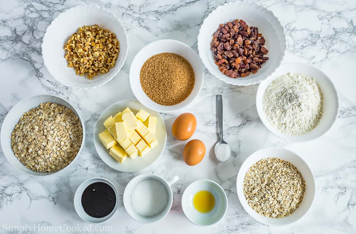 Ingredients for Oatmeal Raisin Cookies, including flour, quick oats, old-fashioned oats, butter, raising, walnuts, eggs, sugar, brown sugar, molasses, baking soda, and salt, on a white background.