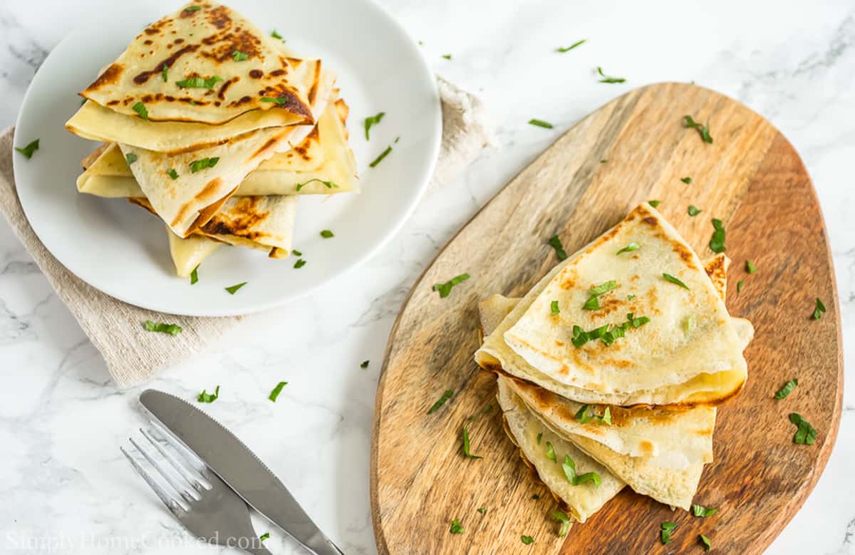 Savory Crepes with Chicken and Mushroom Filling stacked on a wooden board and more on a white plate, with garnish sprinkled on top and a fork and knife nearby.