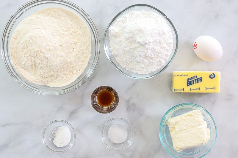 Ingredients for Easy Cream Cheese Cookies, including flour, powdered sugar, butter, cream cheese, vanilla extract, egg, baking powder, and salt, on a white background.