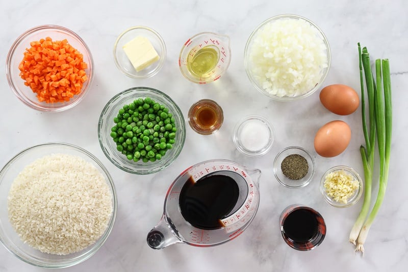 Ingredients for Hibachi Fried Rice, including rice, carrots, onion, garlic, butter, frozen peas, eggs, green onion, soy sauce, teriyaki sauce, sesame oil, salt, and pepper, on a white background.