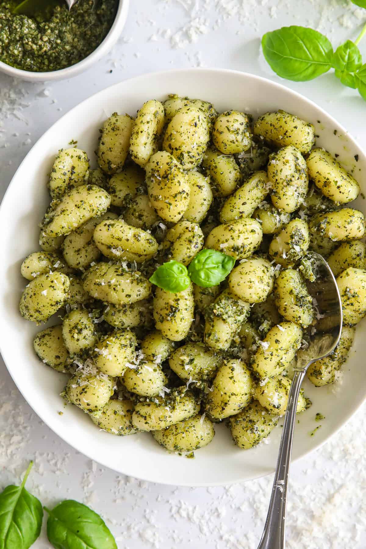 A bowl of Pest Gnocchi with a spoon, and basil, Parmesan cheese, and basil pesto on the side.