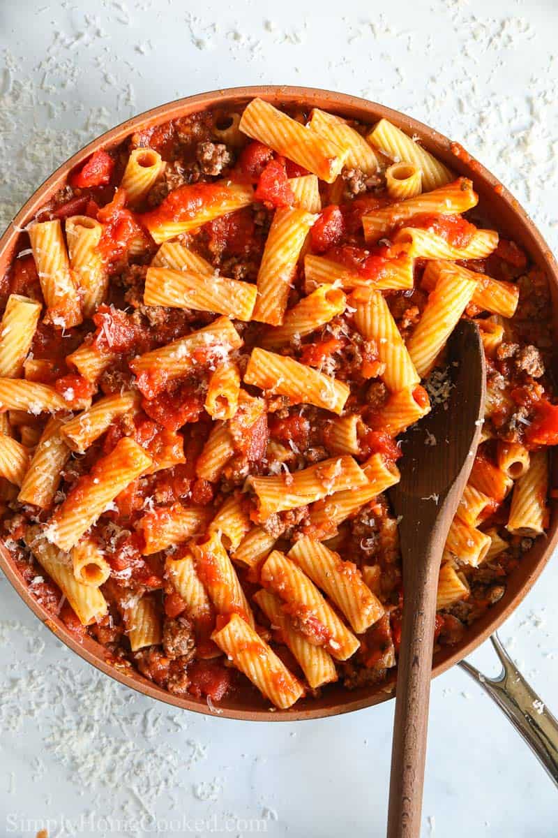 Close up of a skillet full of Rigatoni Bolognese, sprinkled with Parmesan cheese, and with a wooden spoon in it.