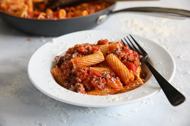 Side view of Rigatoni Bolognese on a plate with a fork, a skillet in the background.