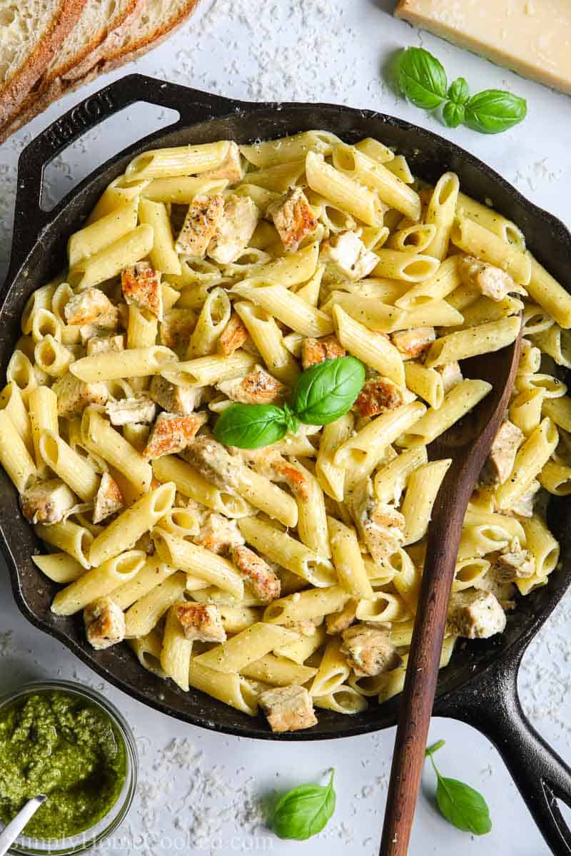 Skillet with Creamy Chicken Pesto Pasta and a wooden spoon in it, and pesto, basil and bread to the side.