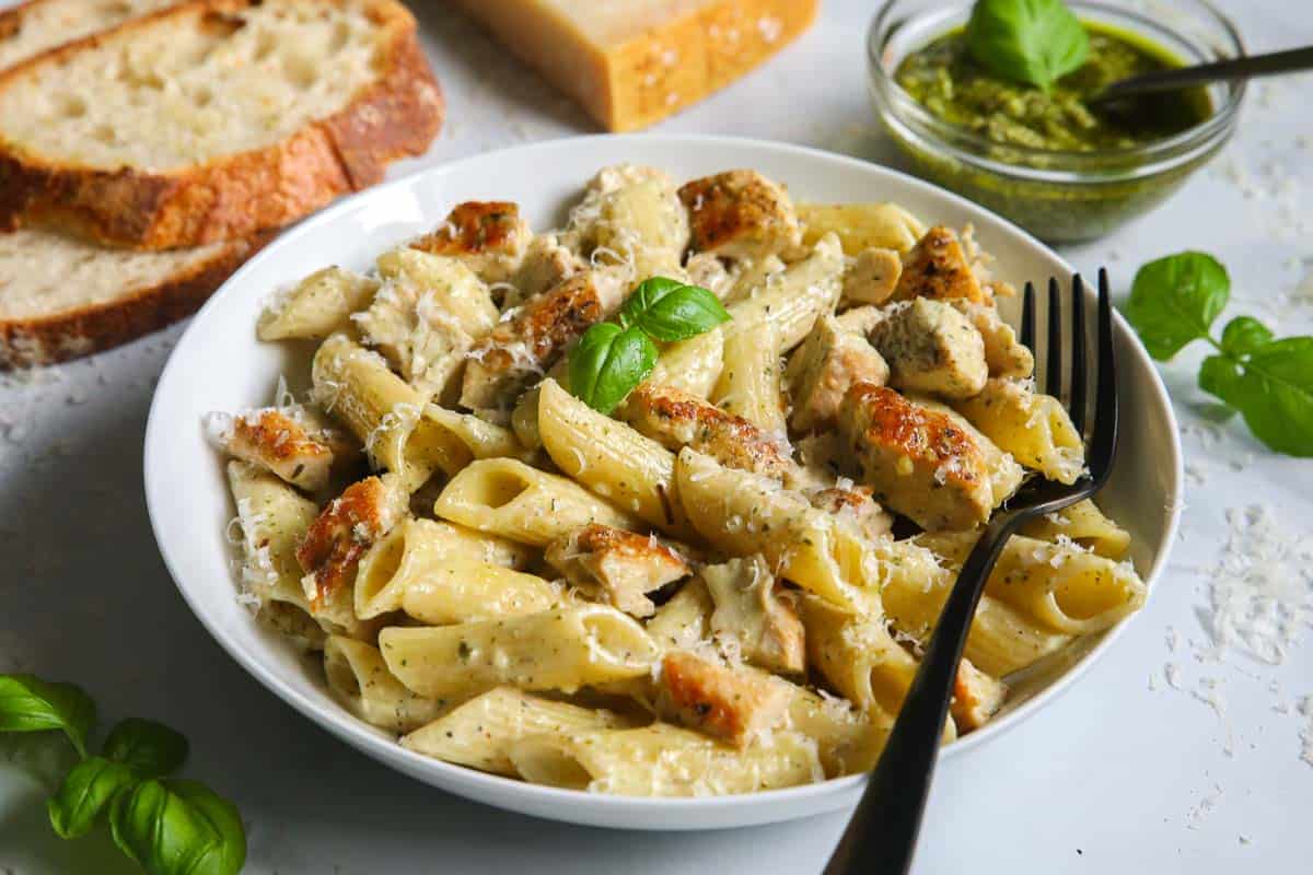 Plate of Creamy Chicken Pesto Pasta with a fork in it and some bread and pesto to the side.
