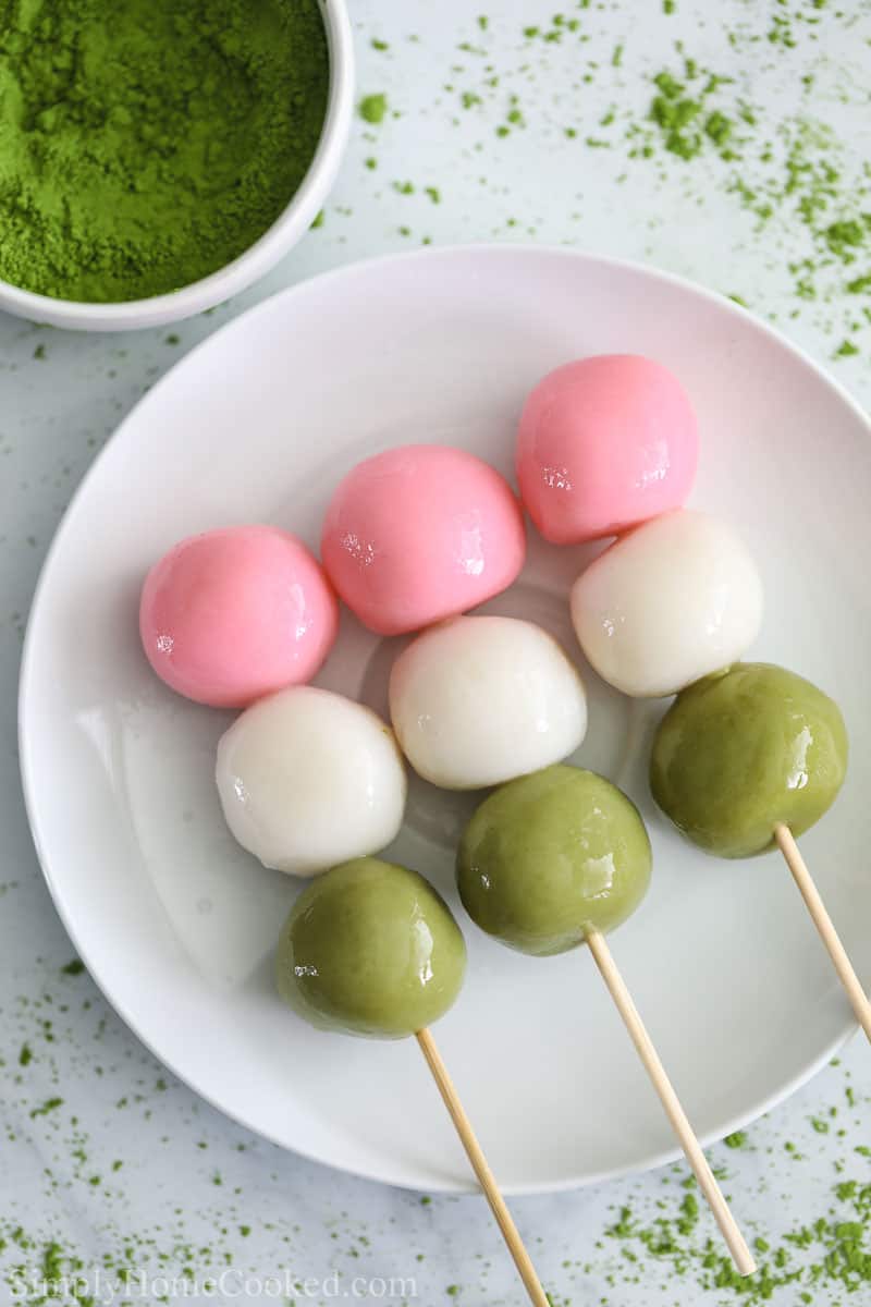 3 Hanami Dango on skewers on a white plate with matcha powder nearby.