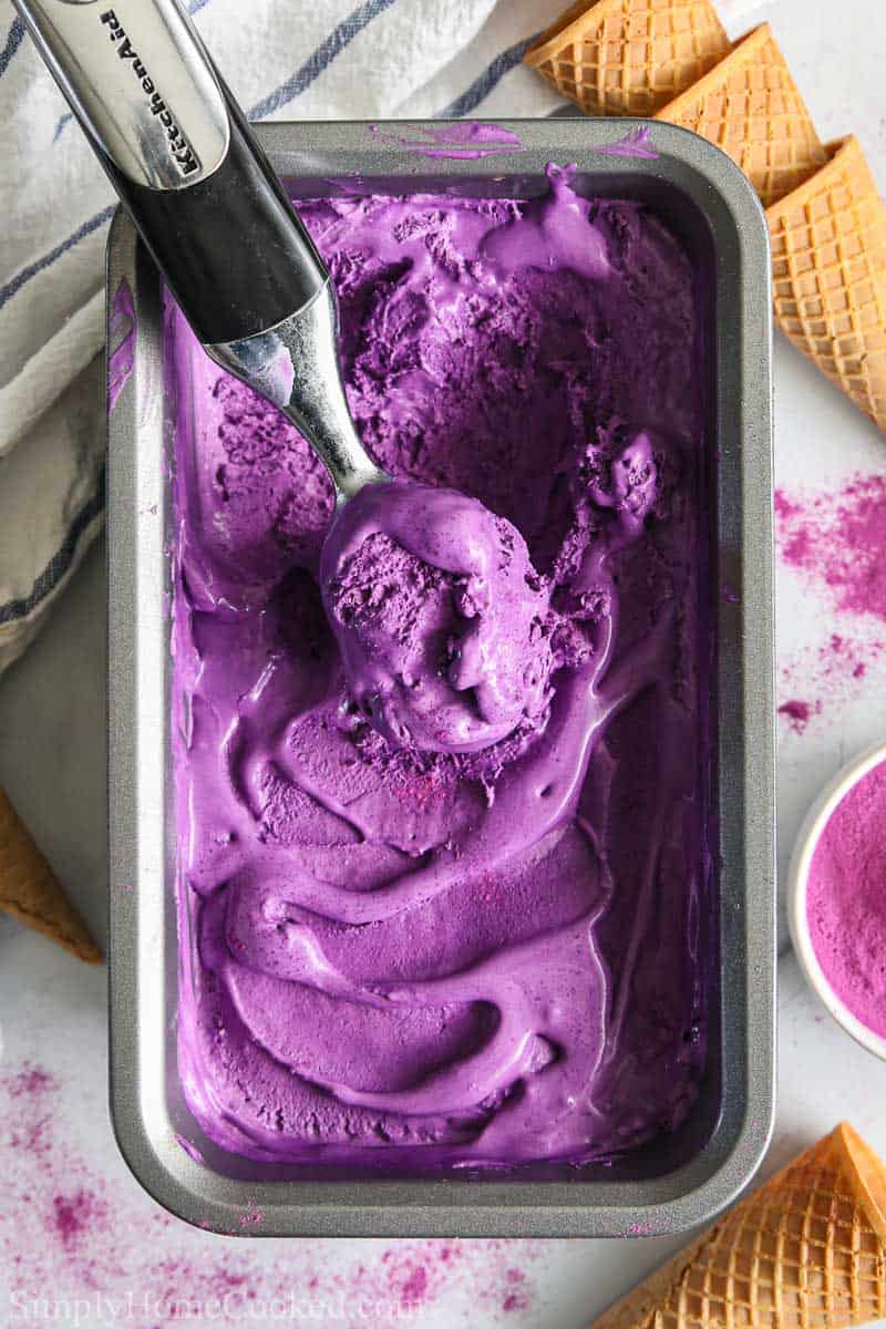 Loaf pan of Easy Ube Ice Cream with an ice cream scooper in the middle, ice cream cones nearby.