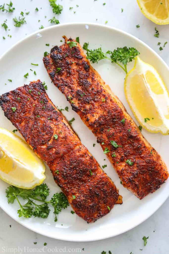 overhead image of two salmon fillets on a white plate with chopped parsley and lemon wedges beside it
