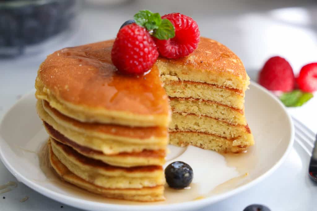 Stack of Almond Flour Pancakes with raspberries, blueberries, and mint on top with syrup, a slice missing.