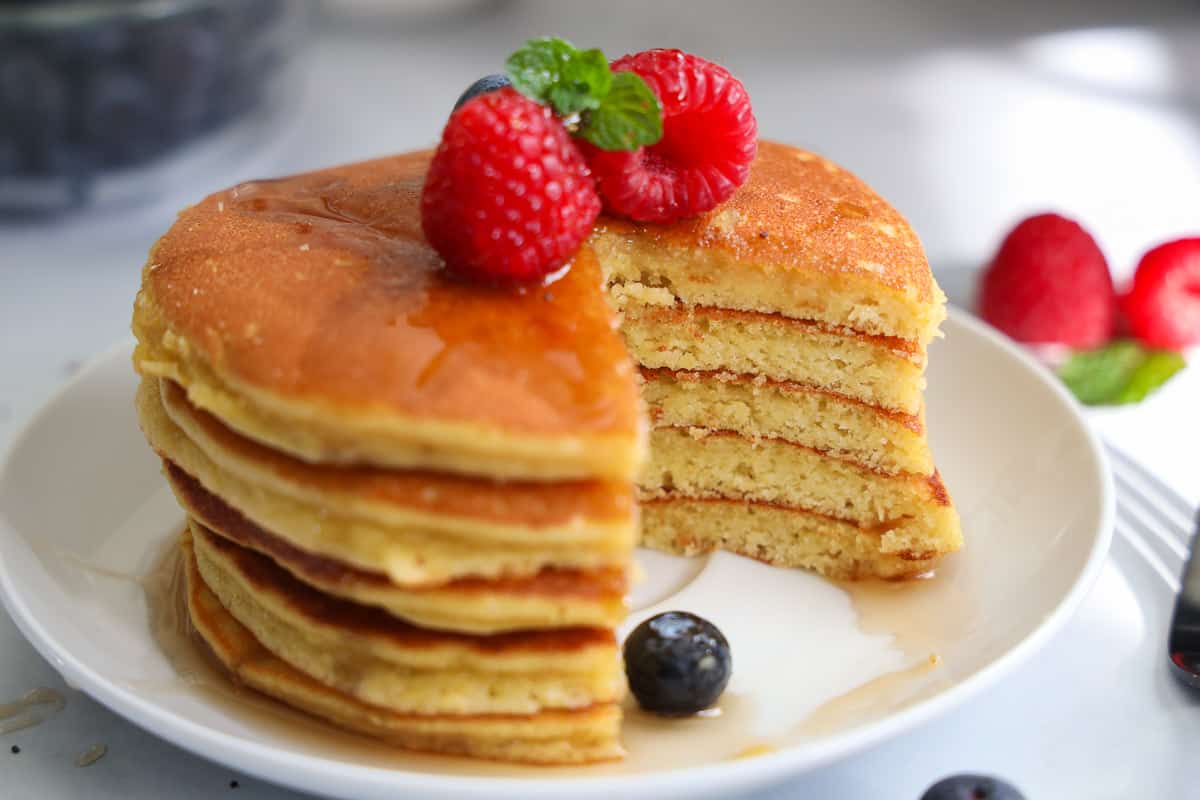 Stack of Almond Flour Pancakes with raspberries, blueberries, and mint on top with syrup, a slice missing.