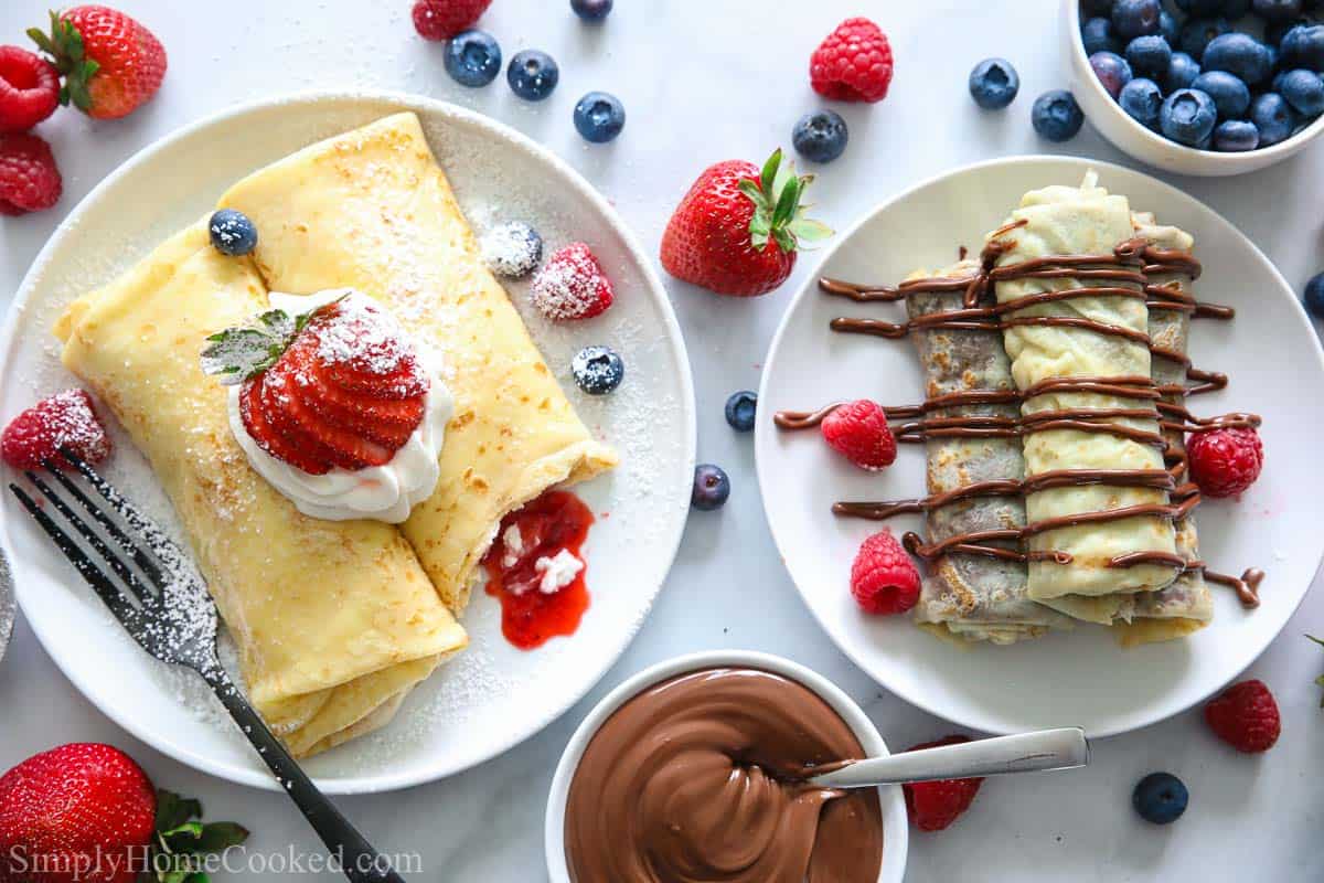 Plates of crepes with strawberries and whipped cream, and drizzled with chocolate sauce, more chocolate and berries nearby