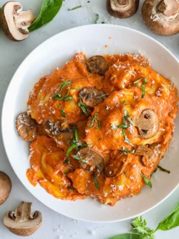 overhead image of sausage ravioli in a white plate with mushrooms and basil beside it
