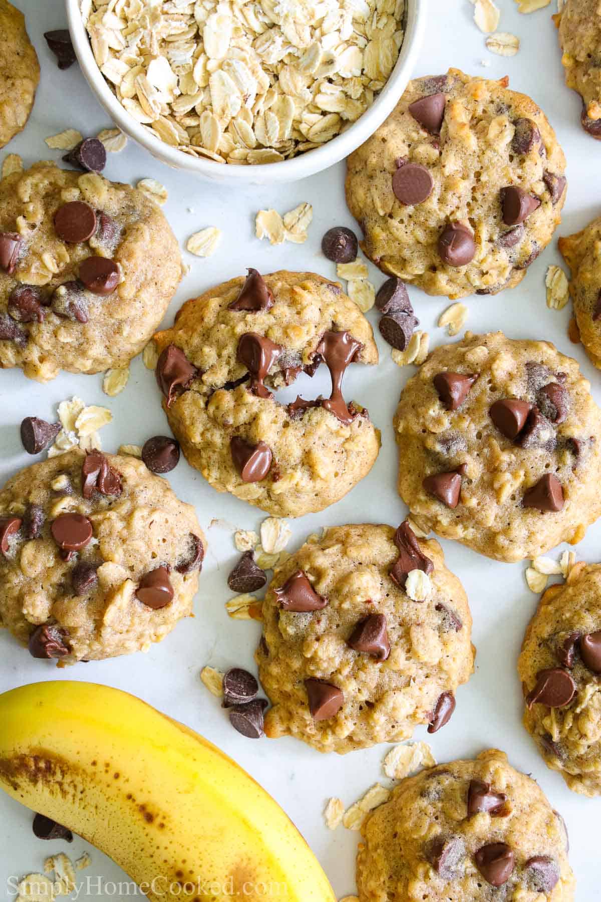 overhead image of banana oatmeal cookies with chocolate chips on top and a bowl of oatmeal and a banana beside them