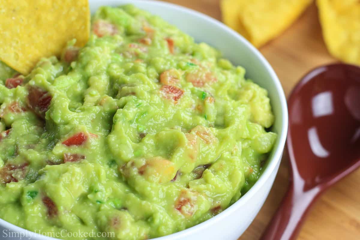 Close up of a chip in a bowl of guacamole with a spoon on the side