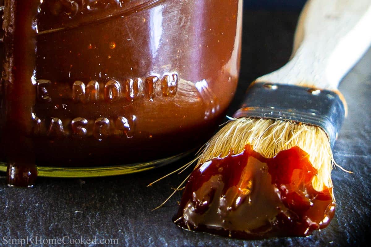 Closeup of mason jar with barbecue sauce and brush next to it, with a dark background.