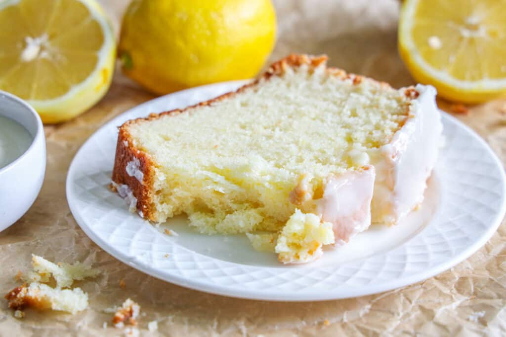 close up image of a slice of lemon loaf on a white plate with lemons in the background