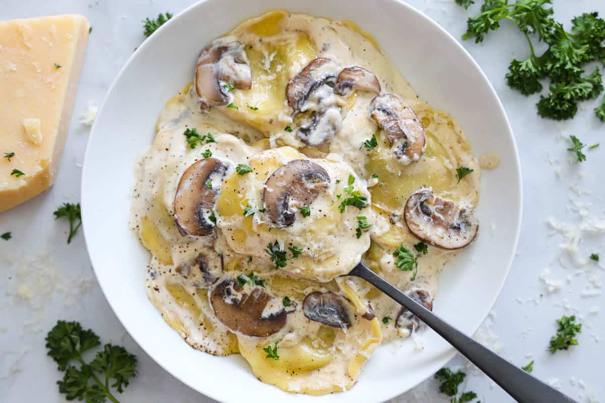 Horizontal image of a plate of Mushroom Ravioli with a spoon, and Parmesan cheese and parsley nearby