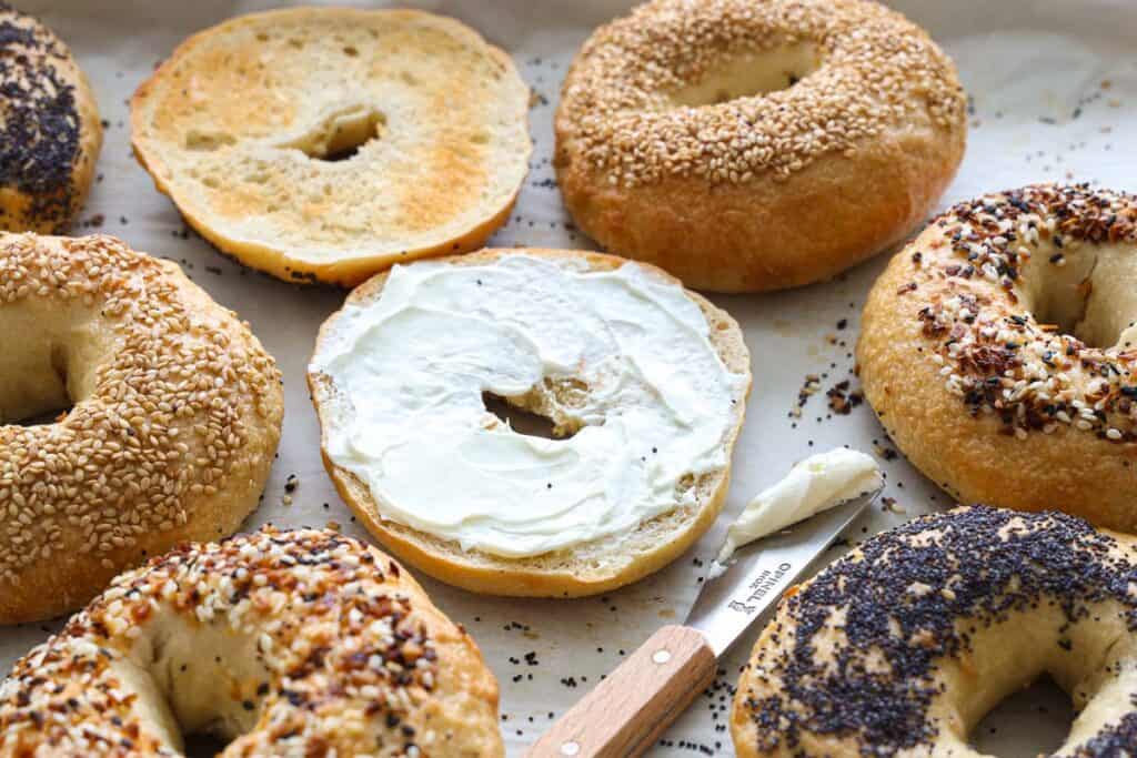 Sourdough bagels with different toppings, one spread with cream cheese, and a knife.
