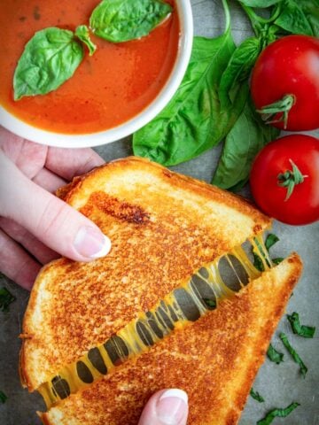 Vertical image of a Grilled Cheese Sandwich being pulled apart, with tomatoes and tomato soup in the background