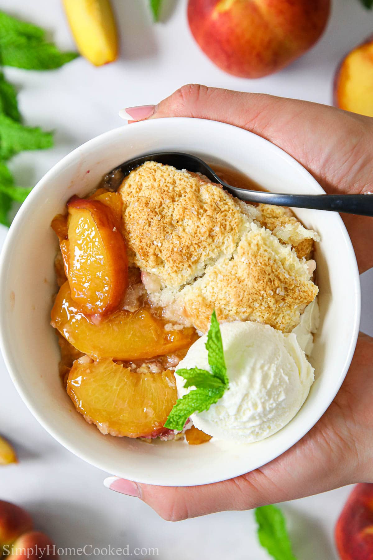Hands holding a bowl of Easy Peach Cobbler with ice cream and a spoon.