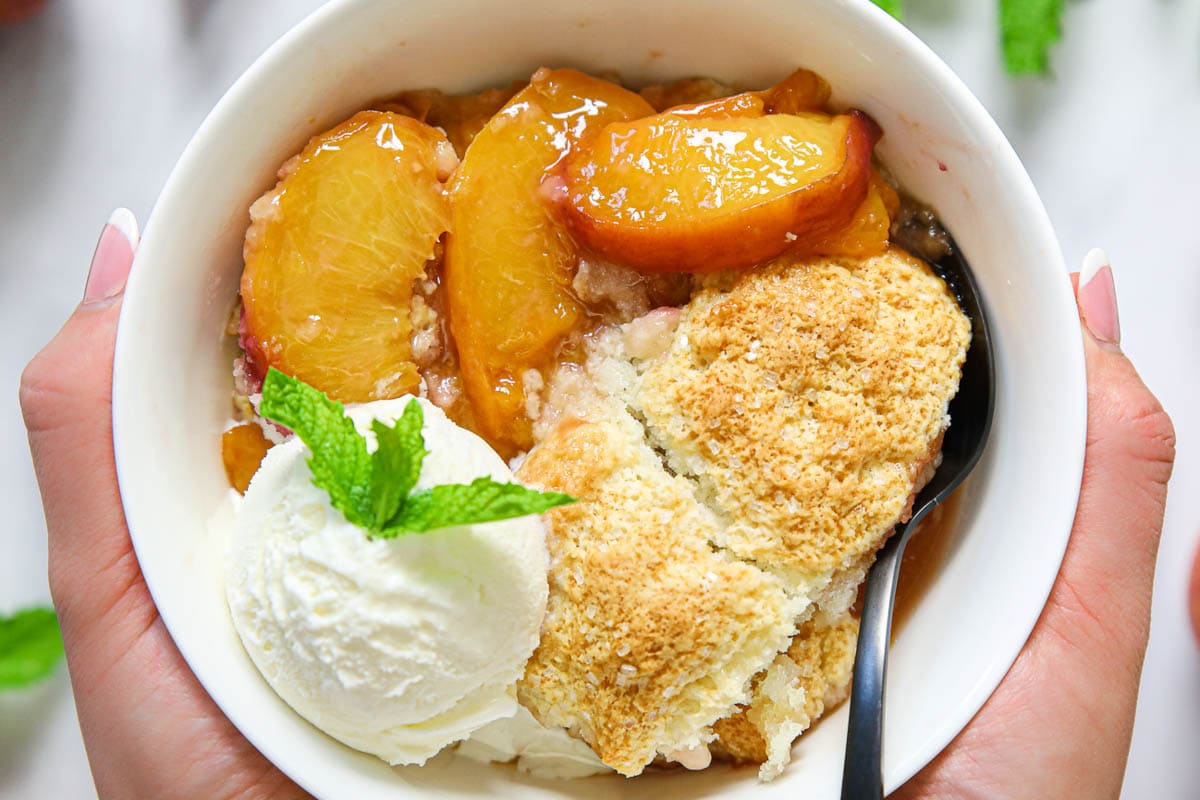 Horizontal image of hands holding a bowl of Easy Peach Cobbler with ice cream and a spoon.