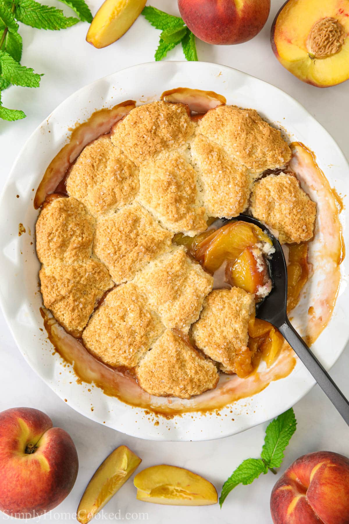 Easy Peach Cobbler in a bowl with a spoon in it and peaches nearby.