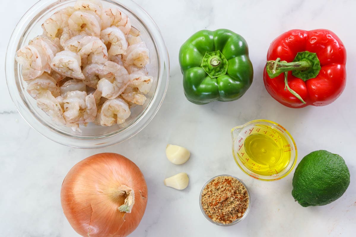 Ingredients for Shrimp Fajitas, including raw shrimp, red and green bell peppers, onion, garlic cloves, lime, olive oil, and fajita seasonings.