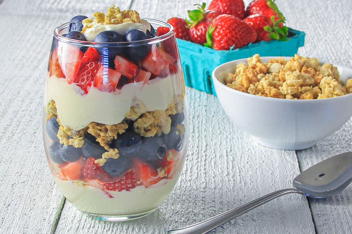 Yogurt Parfait in a glass with a spoon, bowl of granola, and strawberries nearby.