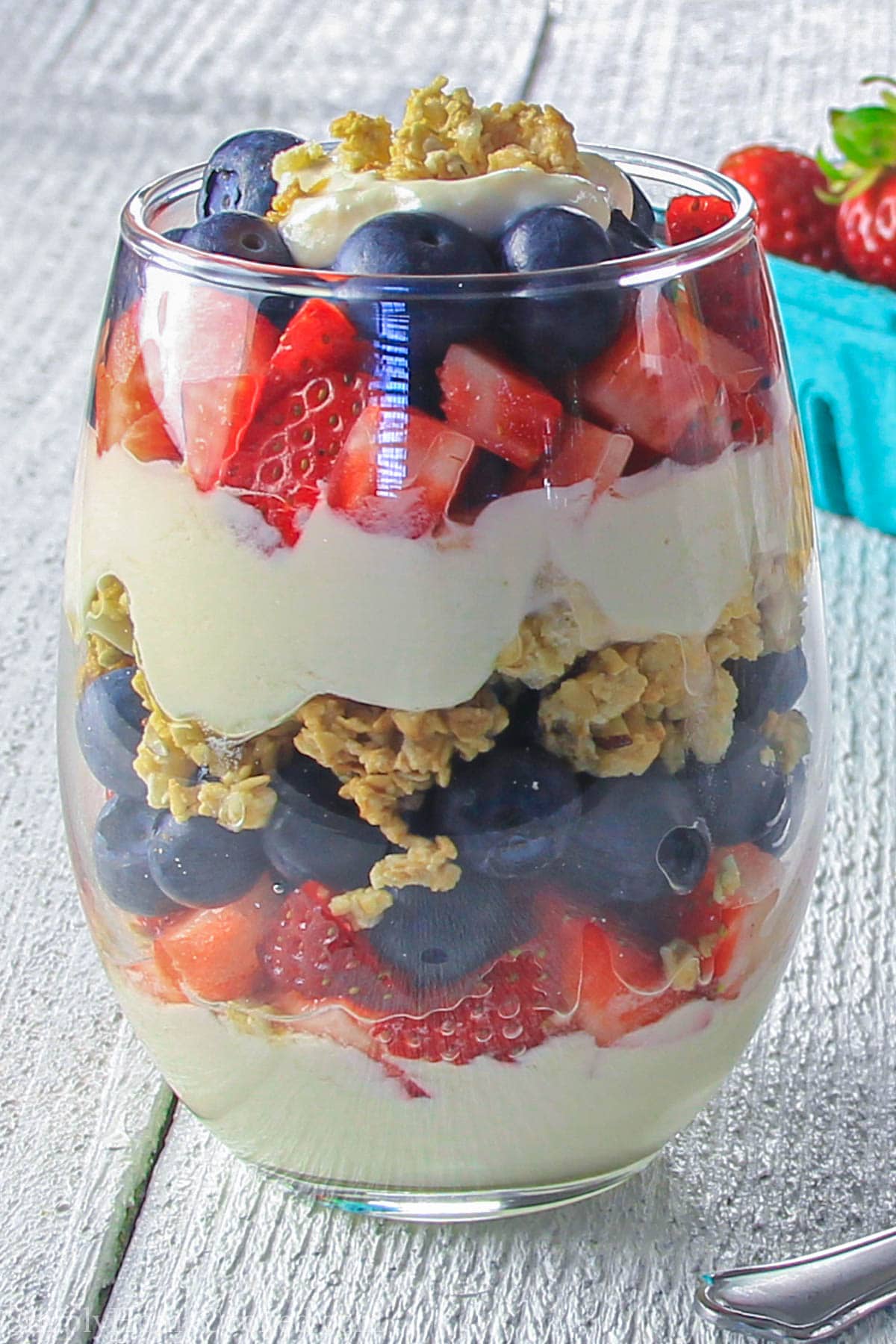 Vertical image of a Yogurt Parfait in a glass, with strawberries in the background.